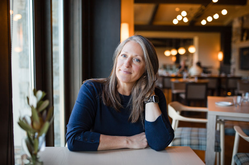 Caryn Gillen, business coach for coaches, seated at a restaurant table with arms resting forward, smiling softly at the camera.
